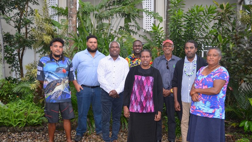 A group of Torres Strait Islanders stand proudly in front of tropical foliage. 