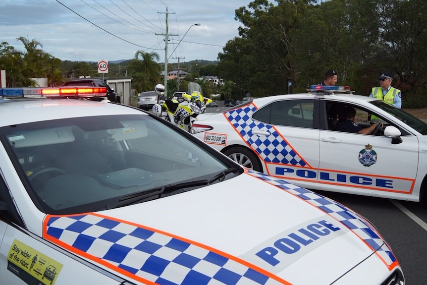 Two police cars with their lights on, a police motorbike and police officers block a suburban street