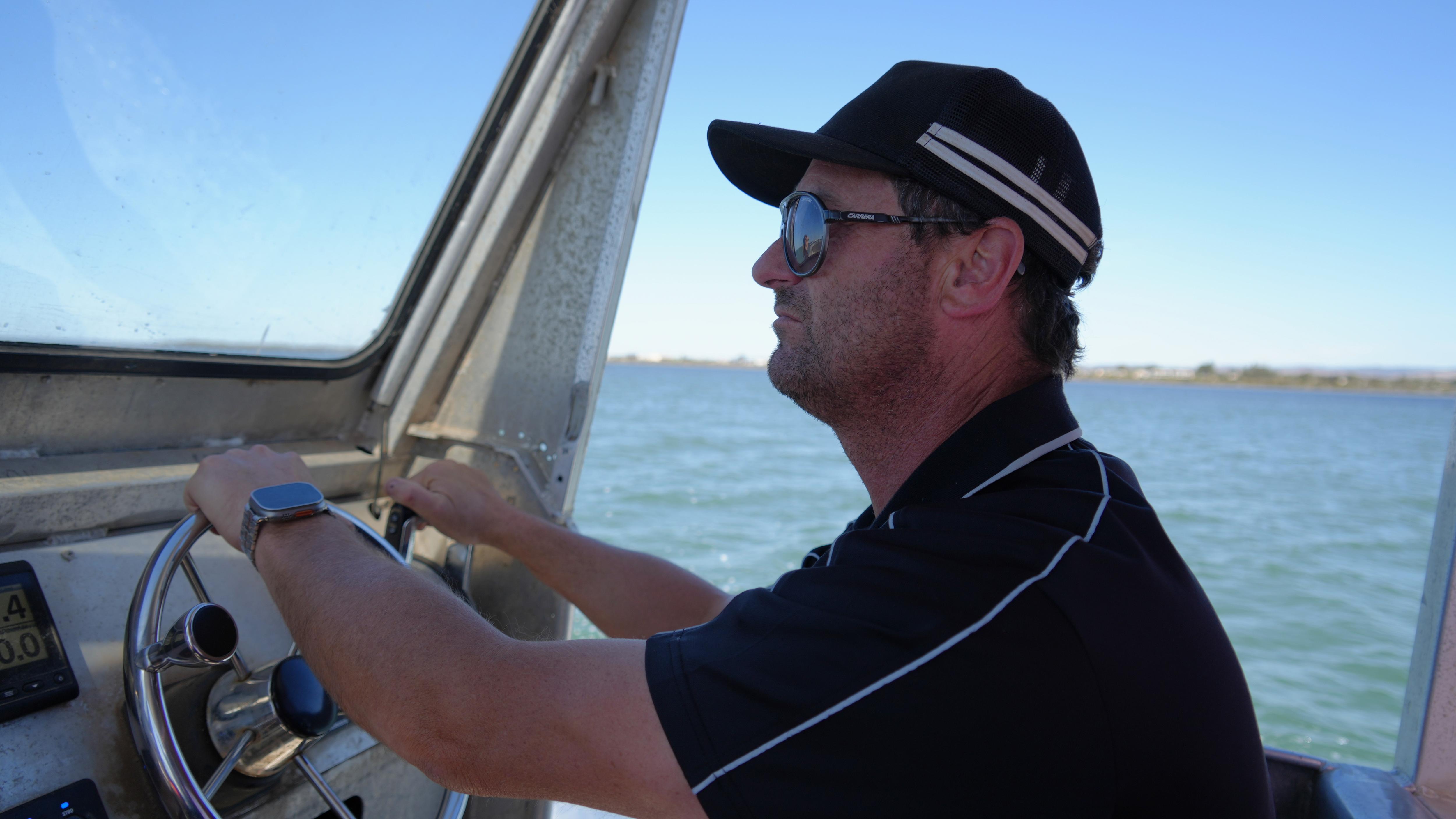 The left-side of a man wearing sunglasses and a cap, holding the steering wheel of a boat, water in the background