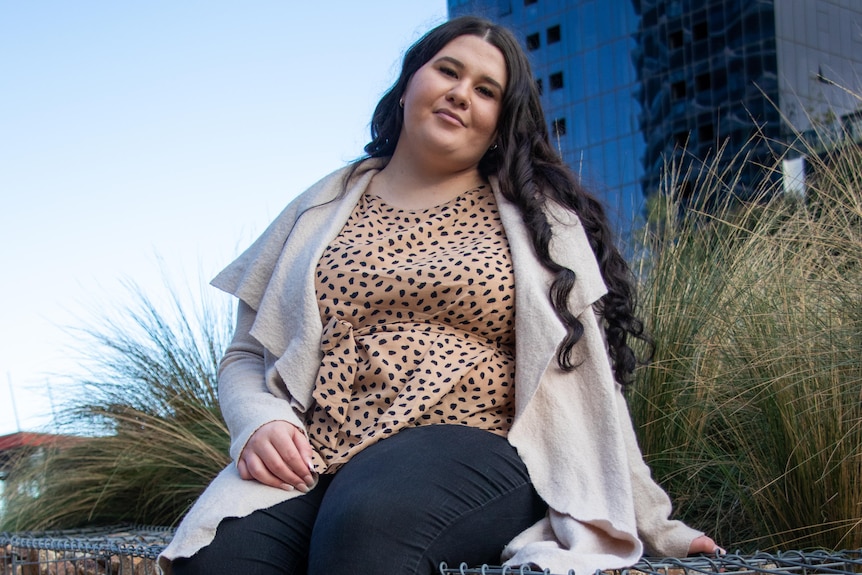 A young woman wearing a leopard print top, beige cardigan and dark jeans sits on a rock wall in front of a skyscraper