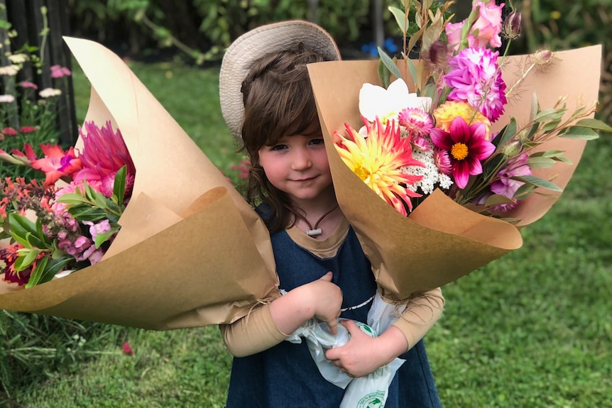 A little girl holds two large bunches of flowers wrapped in brown paper