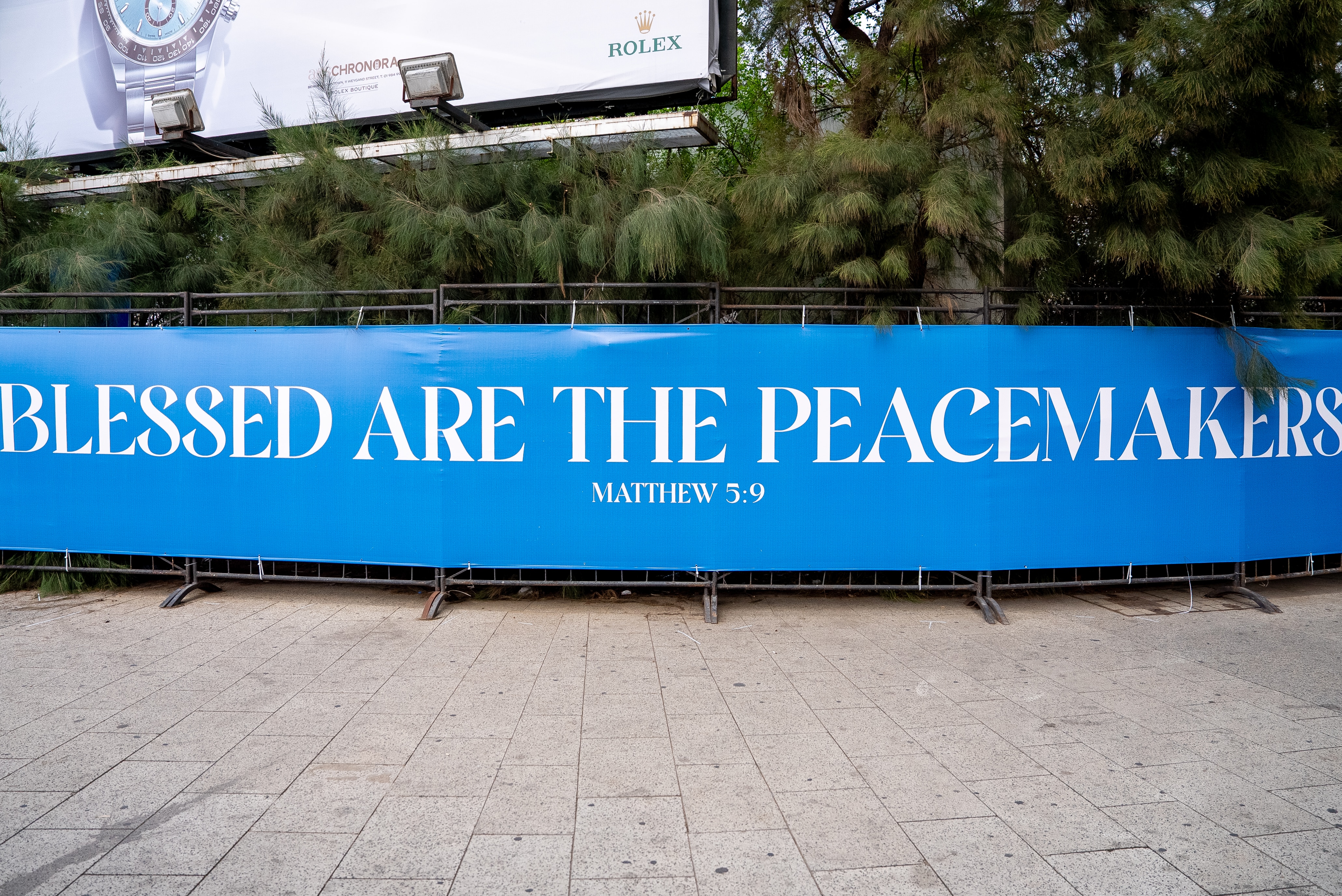 A sign stretched along metal barriers that reads Blessed are the peacemakers Matthew 5:9 in front of some trees.