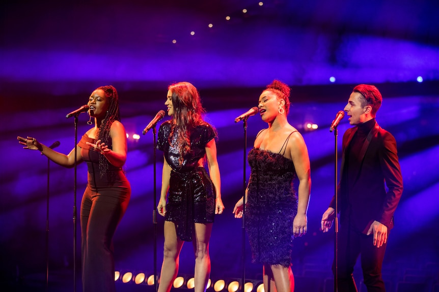 Four performers sing into microphones on a blue-lit stage