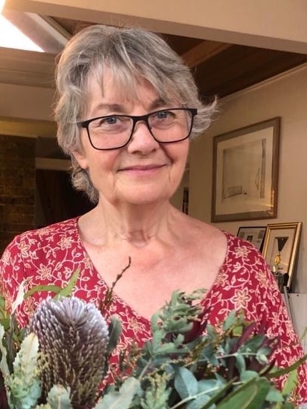 A smiling middle-aged woman, short grey hair, red printed top, wears glasses, native flowers in front, frames on wall behind.