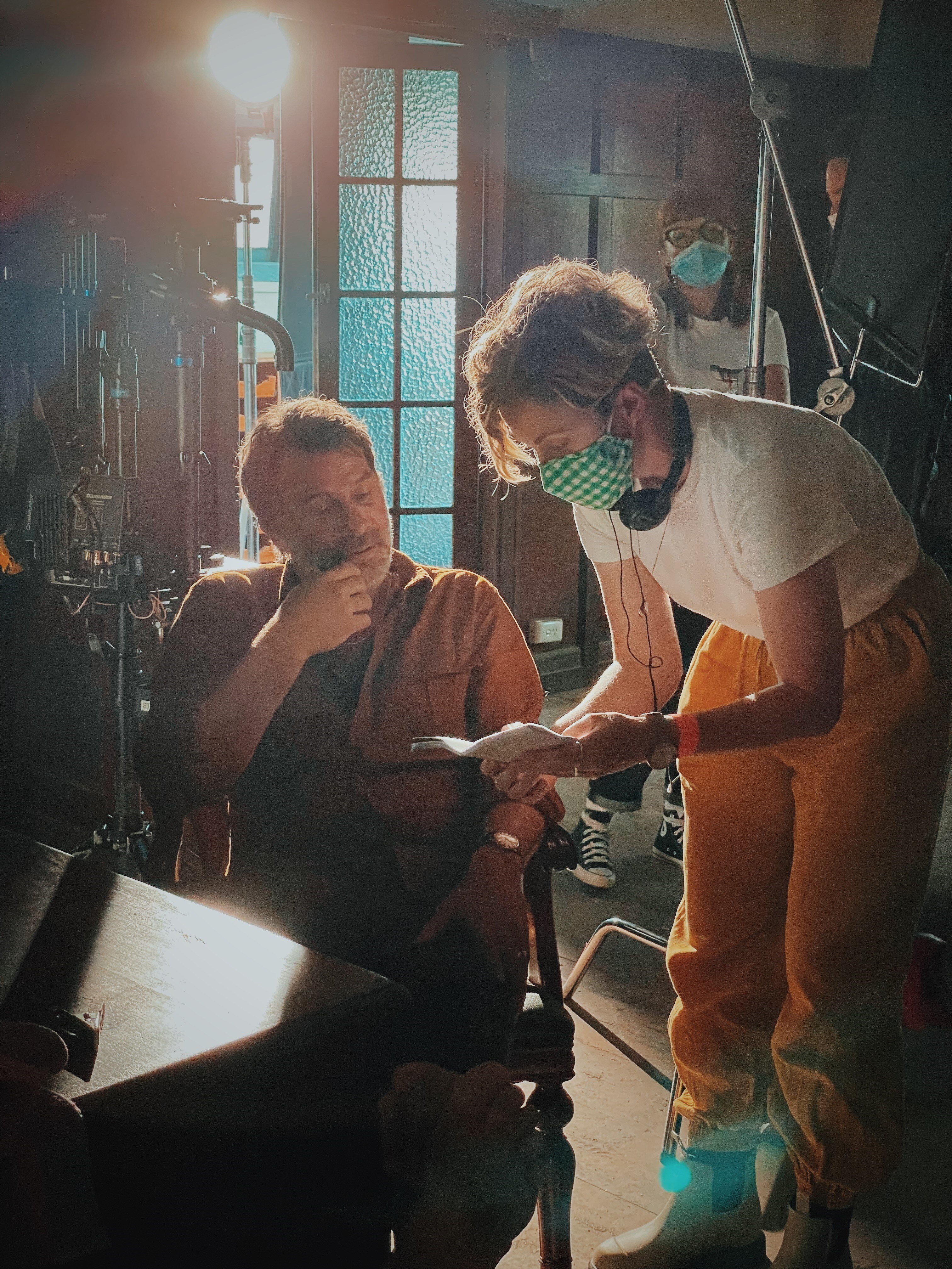A woman wearing a green mask leaning over and talking to a man who is sitting.