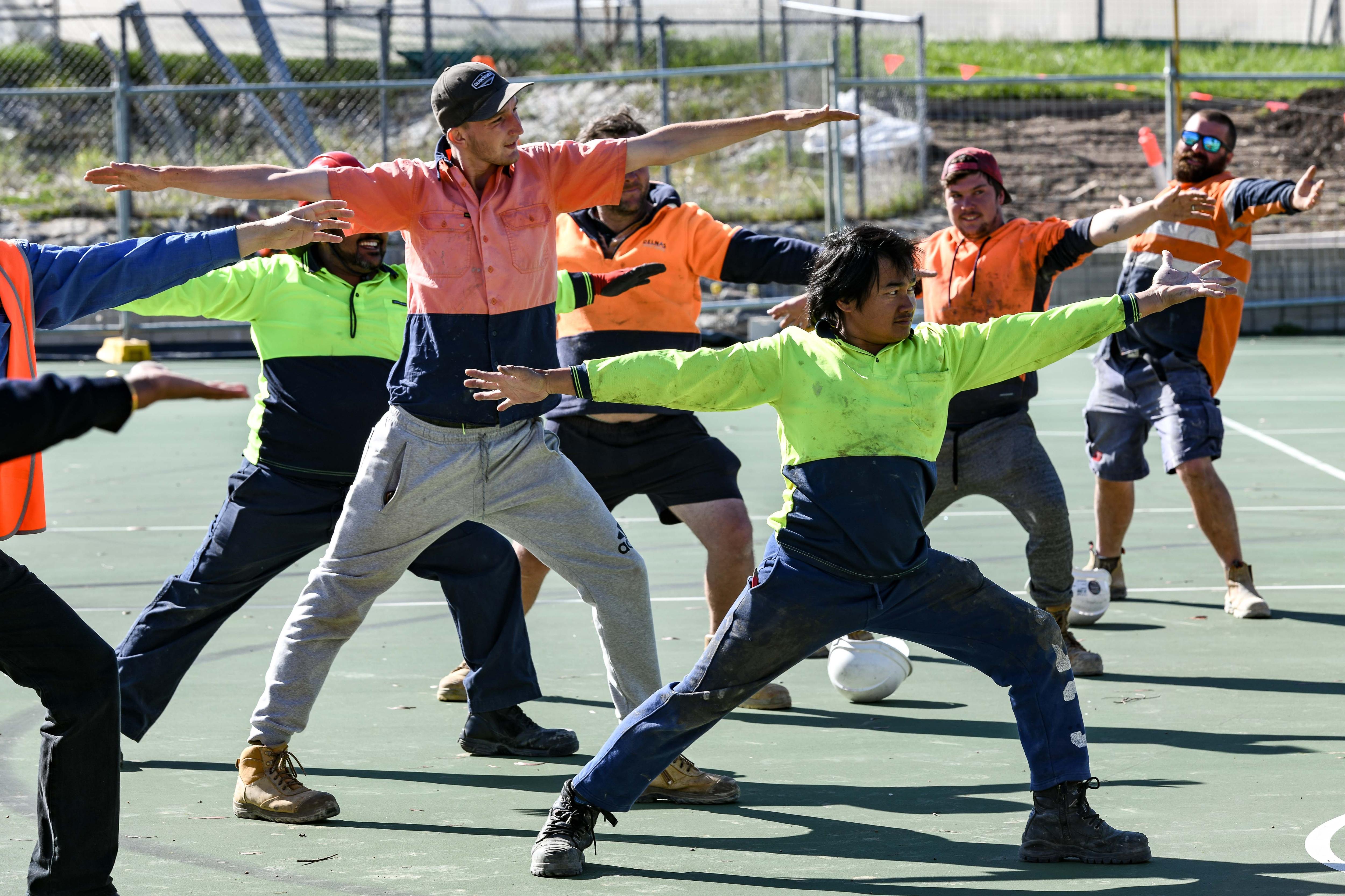 A group of men in high-vis stand in a warrior 2 yoga pose at the construction site.
