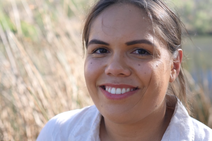 A close up of a young Aboriginal woman smiling into the camera with grasses behind her.