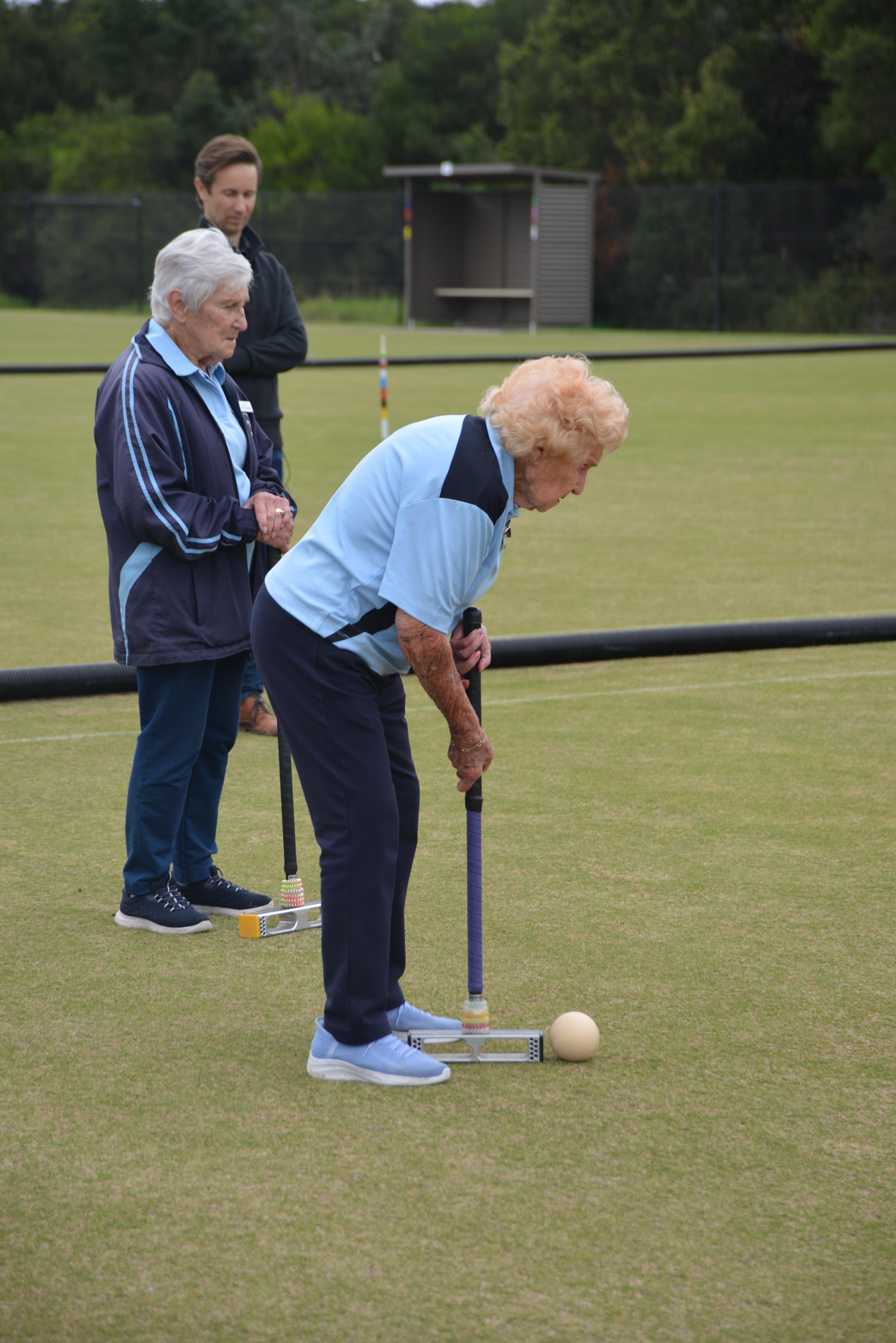 A woman with a blonde perm and wearing a blue sports uniform hits a croquet ball. 