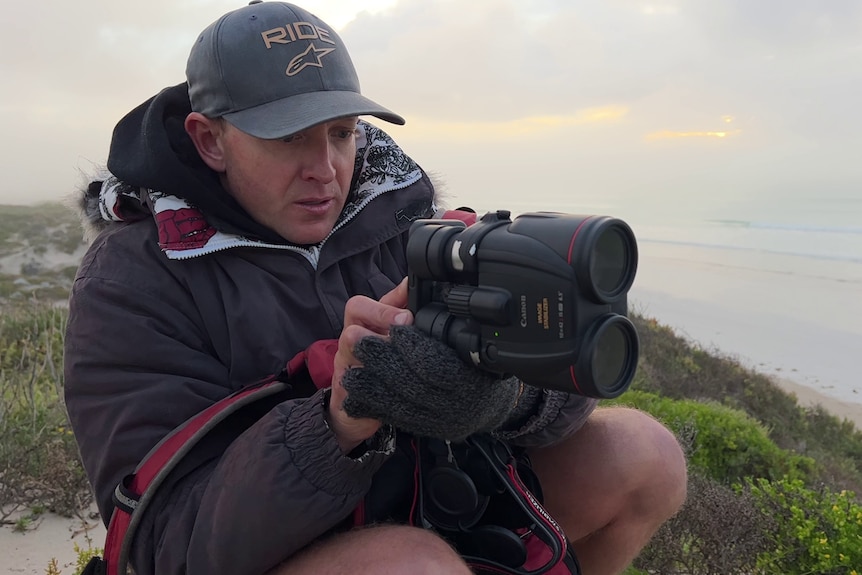 Man wearing coat and cap, holding phone to film through binoculars, sitting on hill with misty ocean background