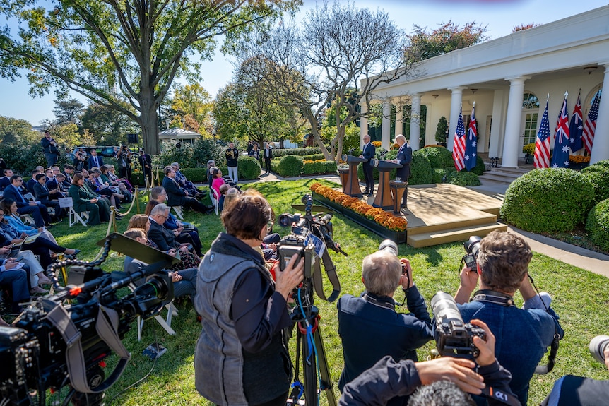 Reporters and photographers surround the two leaders, who are at podiums in front of Australian and US flags in a garden.
