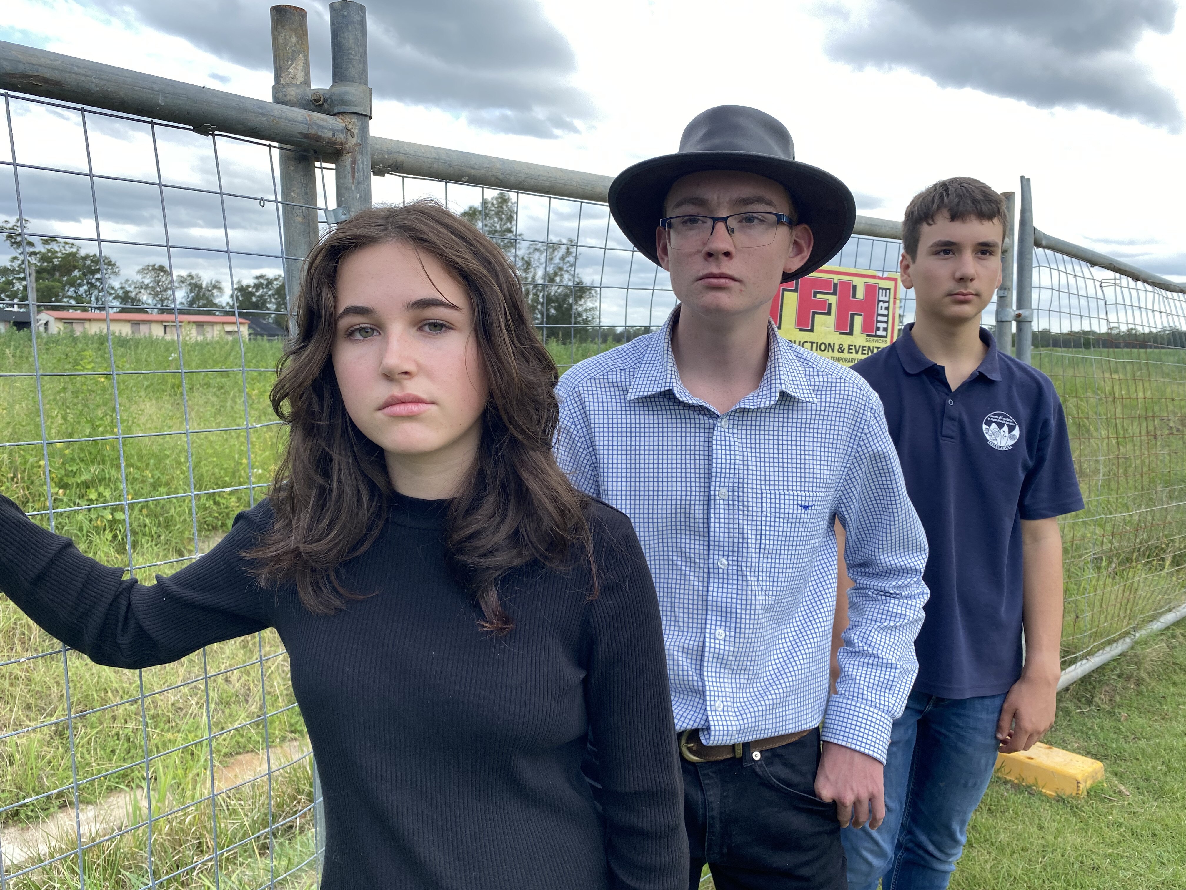 Three students stand in front of a fence watching the empty field where there school is supposed to be. 