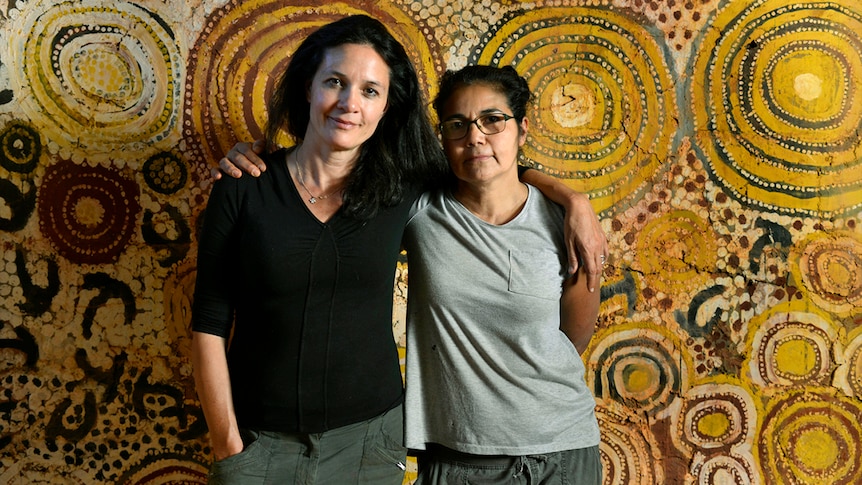 Black and white photo of two women standing against a wall with Indigenous art.