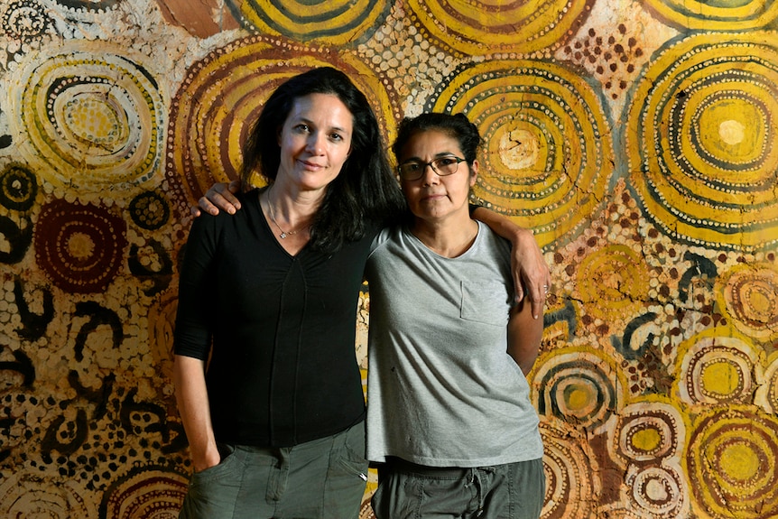 Black and white photo of two women standing against a wall with Indigenous art.