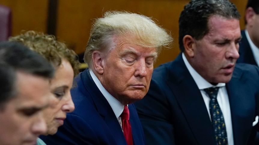 Donald Trump glances down, his face in a tight frown, sitting between two men in suits