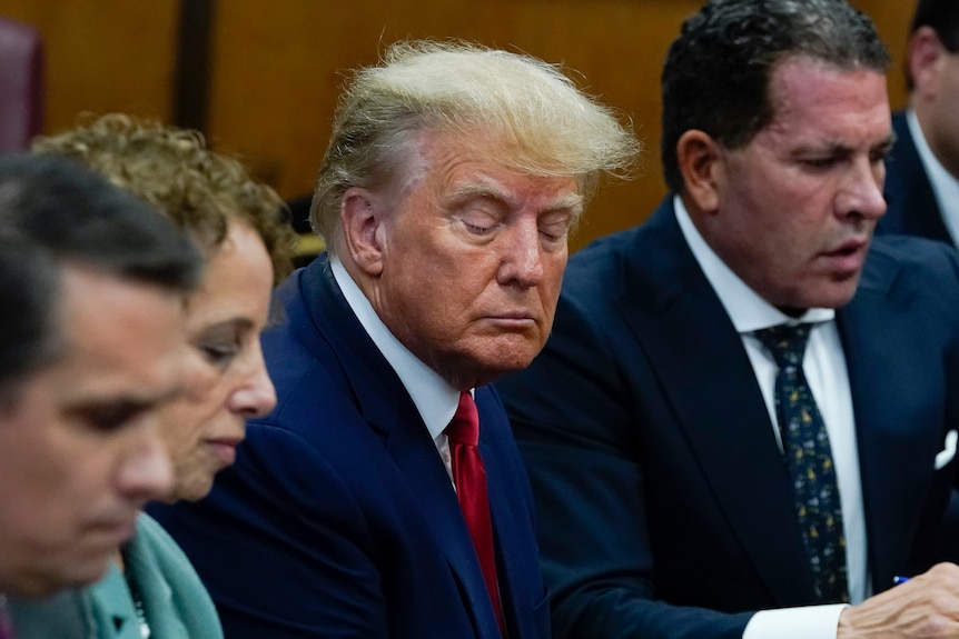 Donald Trump glances down, his face in a tight frown, sitting between two men in suits