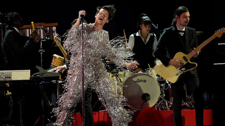 A man in a sparkly, stringy silver suit dances while holding onto a microphone stand onstage at the Grammys.