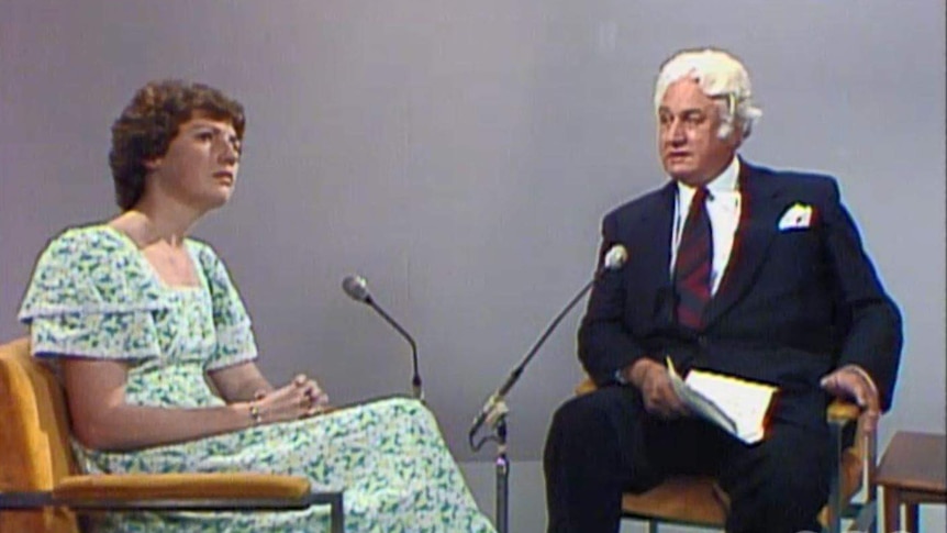 A woman and a man sitting in front of microphones on stage talking for a televised interview.