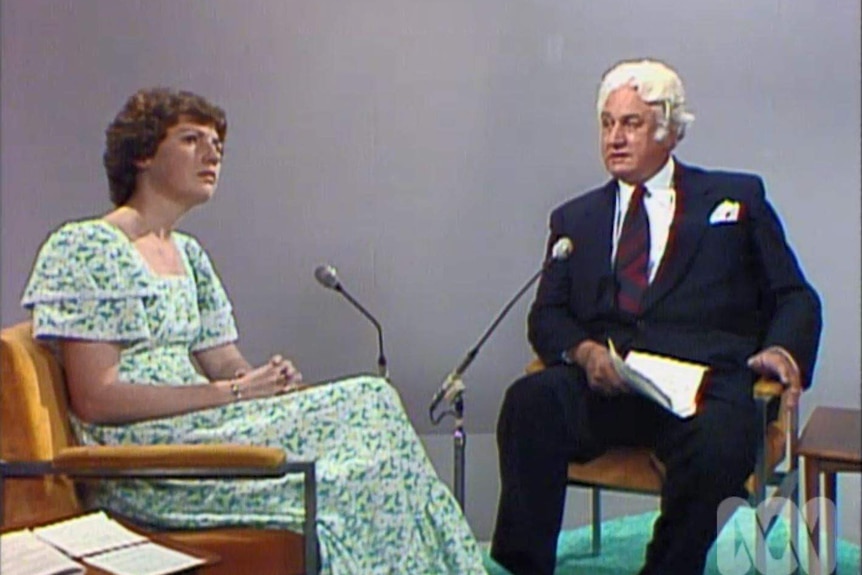 A woman and a man sitting in front of microphones on stage talking for a televised interview.