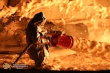 A firefighter in a large hazmat suit uses a large hose to shoot foam towards a fire.