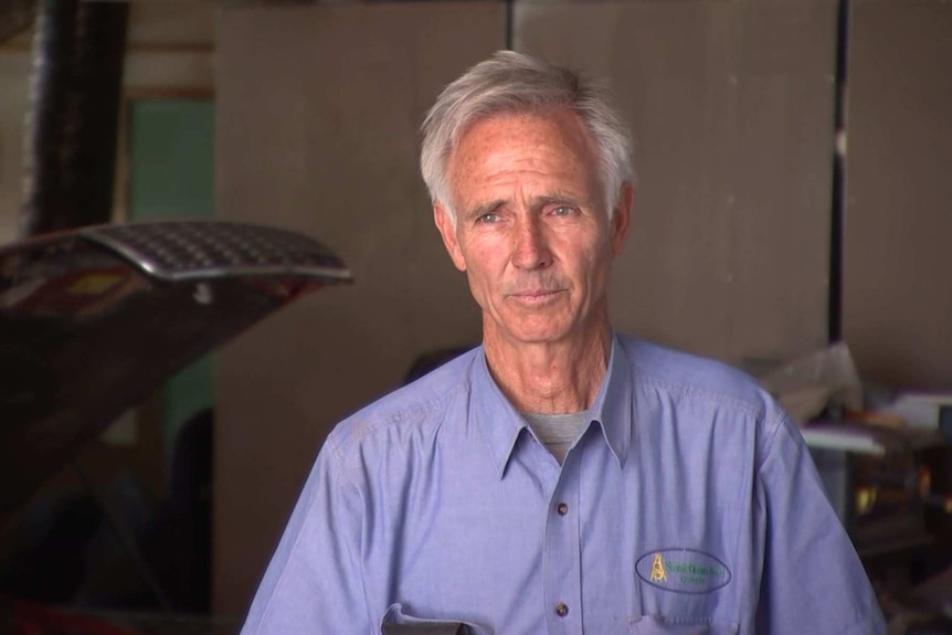 Man  with grey hair looking to the side of camera with a car workshop in the background