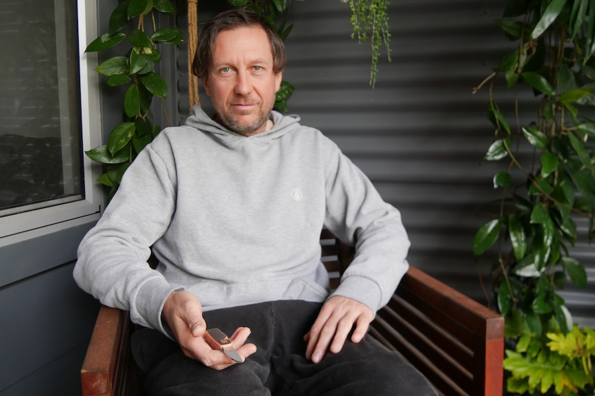 A man holding a pocket knife sitting on a chair in front of a corrugated wall