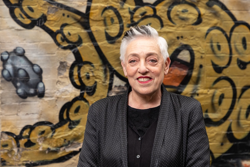Middle-aged white woman with short white hair wears a black jacket over a black blouse standing against a graffitied wall