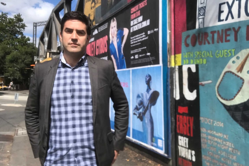 A young man in a grey suit coast and checked shirti stands on the street, in front of  posters for music gigs.