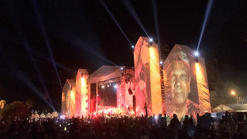 Hundreds of people sit in front of a large stage lit in red and orange with a large image of an Aboriginal man.