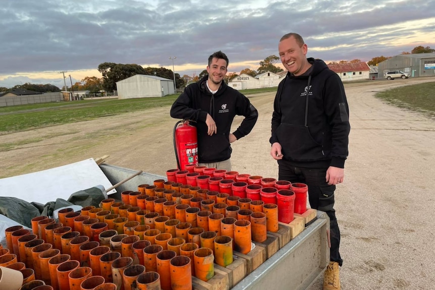 Two men stand behind a trailer of fireworks mortar tubes.