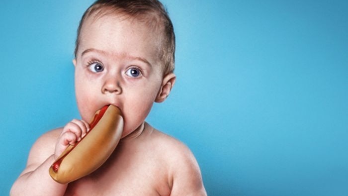 A toddler in a nappy bites a hotdog while a plate of burgers sits nearby.