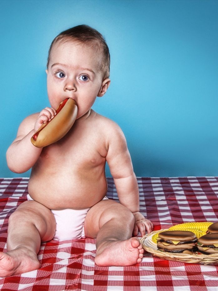 A baby wearing a nappy sits on a red and white tablecloth eating a hot dog next to a plate of burgers.
