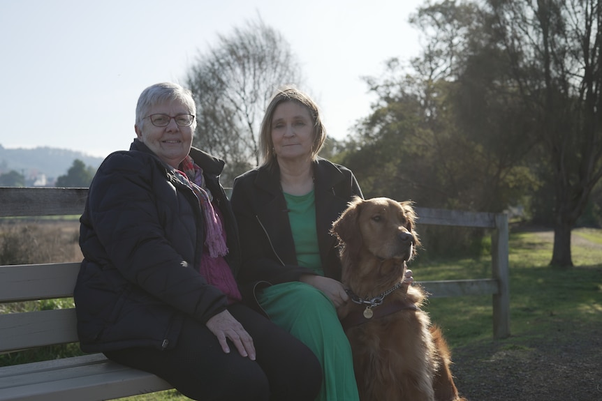 Two women sitting on a park bench. One woman holds onto a guide dog.