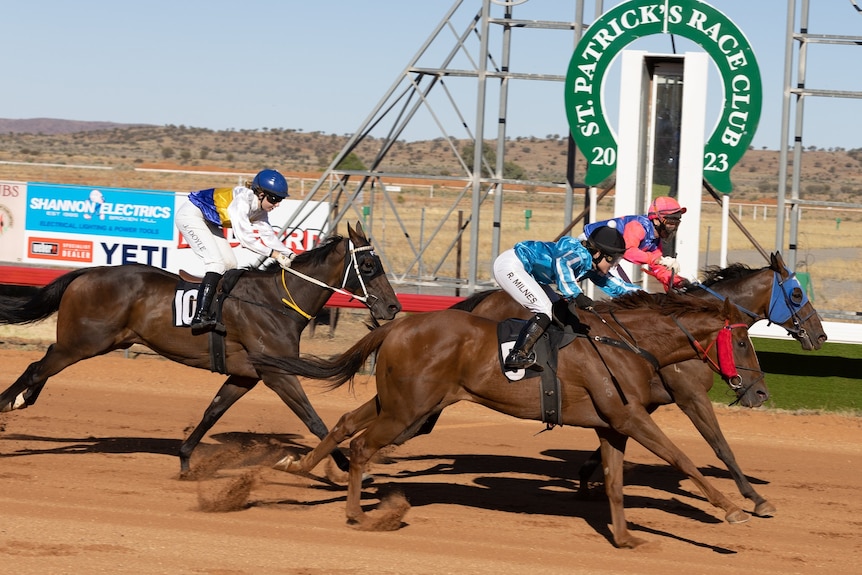 three horses running on a dirt track towards the finish line