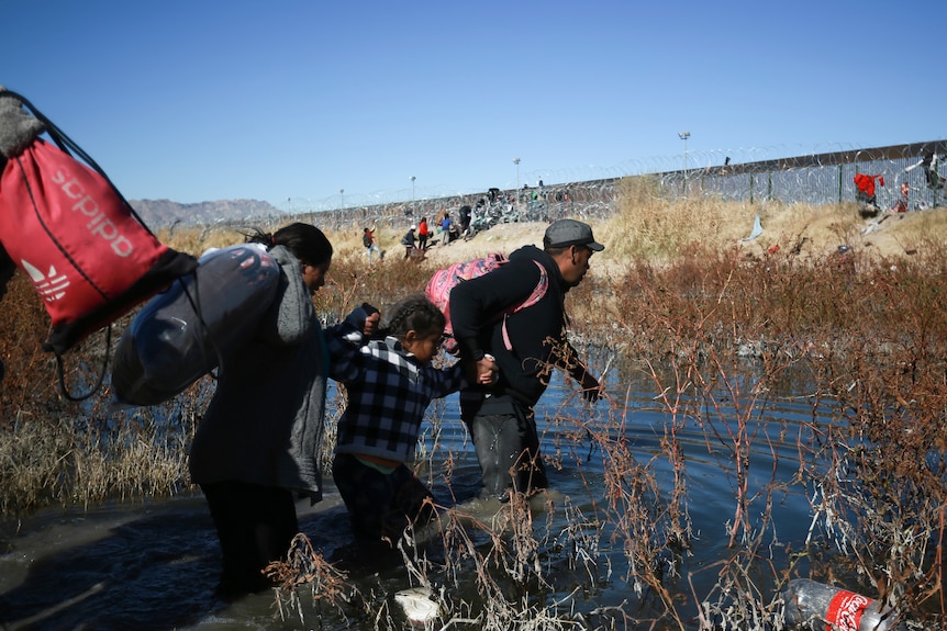 A man, woman and child carry luggage as they wade through knee-deep water in front of a barbed wire fence.