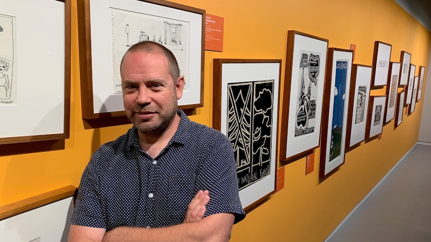 A man stands in front of linocut posters hanging on a yellow wall in an art gallery.