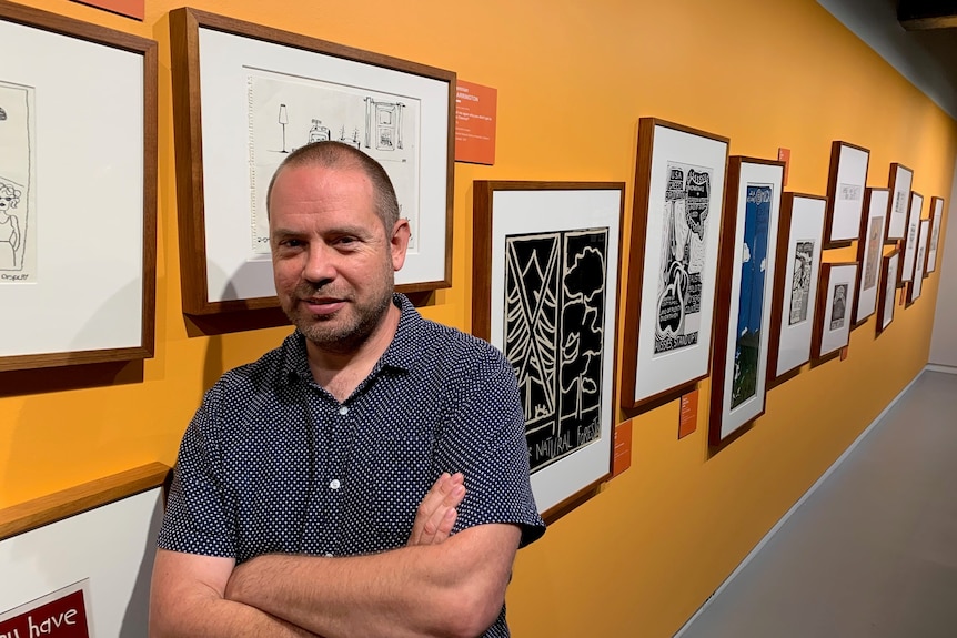 A man stands in front of linocut posters hanging on a yellow wall in an art gallery.