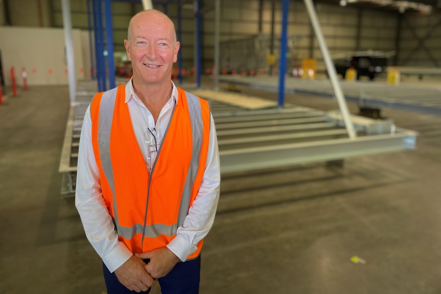 a man stands in an industrial-looking warehouse wearing an orange high-vis vest over his shirt, smiling