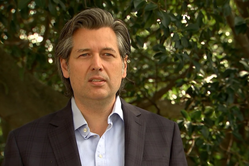 A man in a dark blazer with a light shirt stands under a tree.