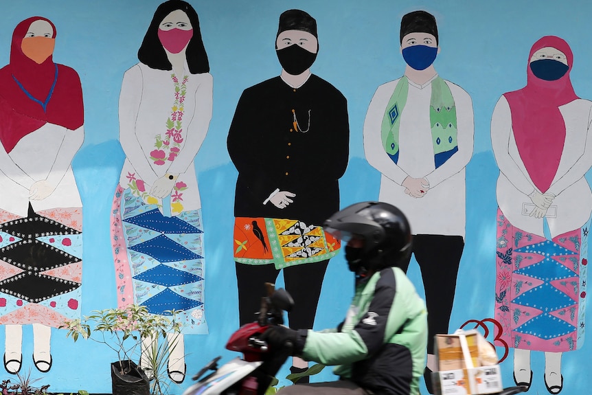 A motorbike rides past a mural depicting people in traditional Indonesian attire wearing face masks