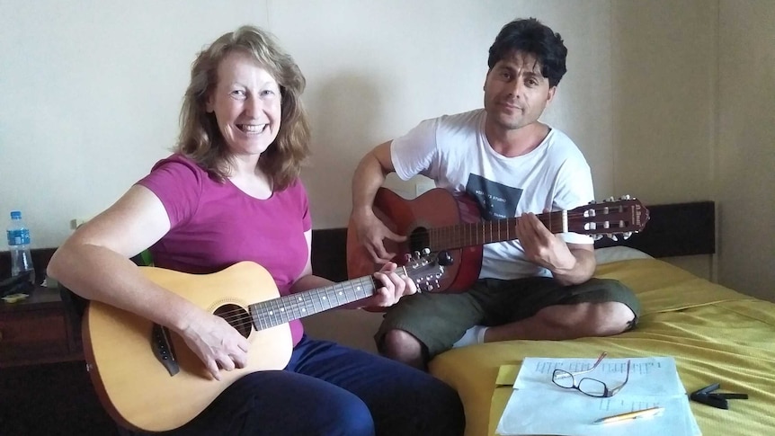 Australian woman with refugee holding guitars in a hotel room, notebooks on the bed.