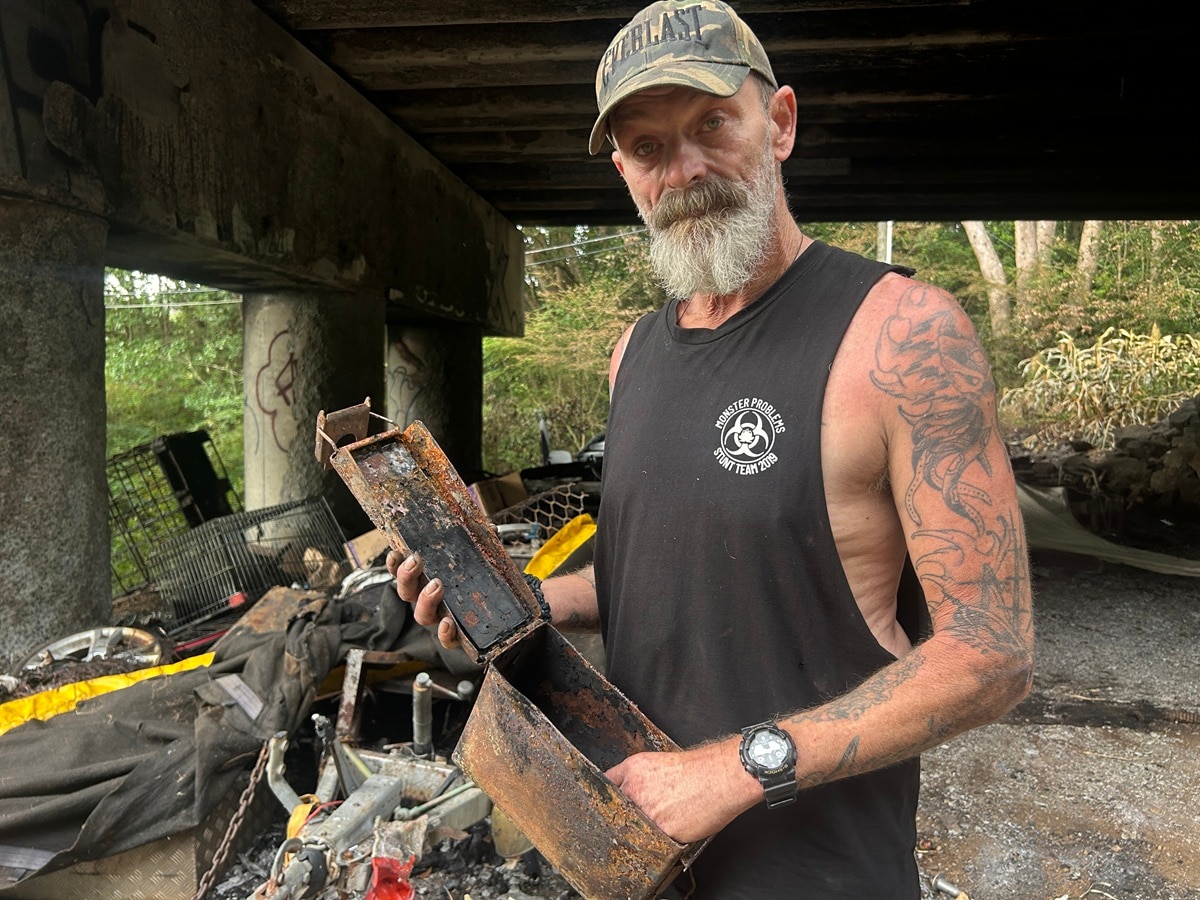 Un hombre se encuentra debajo de un puente, rodeado de cenizas, sosteniendo una caja quemada.