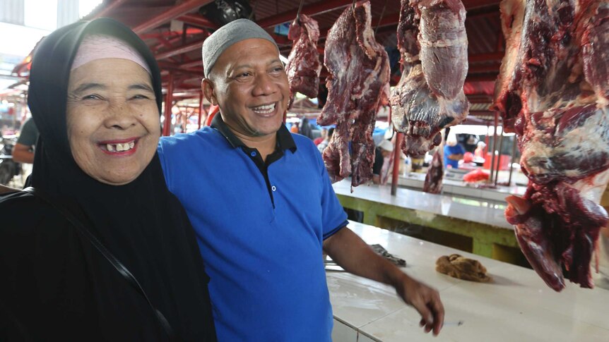 An Indonesian man and woman smile at a market selling fresh meat, hanging on hooks in the background.