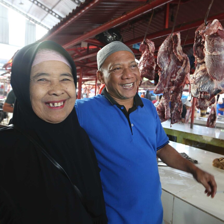 An Indonesian man and woman smile at a market selling fresh meat, hanging on hooks in the background.