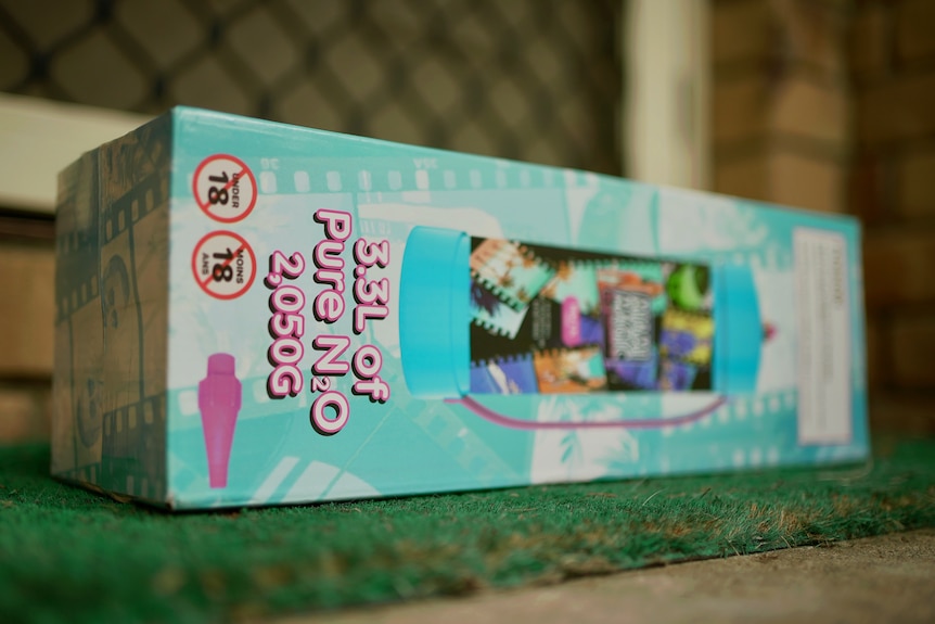 An image of a light blue cardboard box containing nitrous oxide on a welcome mat at a front door with flyscreen in background
