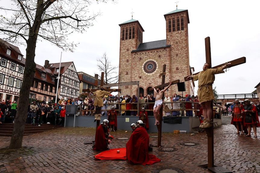 Three men are crucified on a cross in front of a church, with three men dressed as Roman soldiers and a crowd watching on.