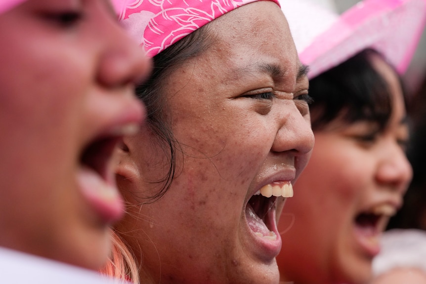 A close up image shows three female activists shout chants while wearing pink headware.