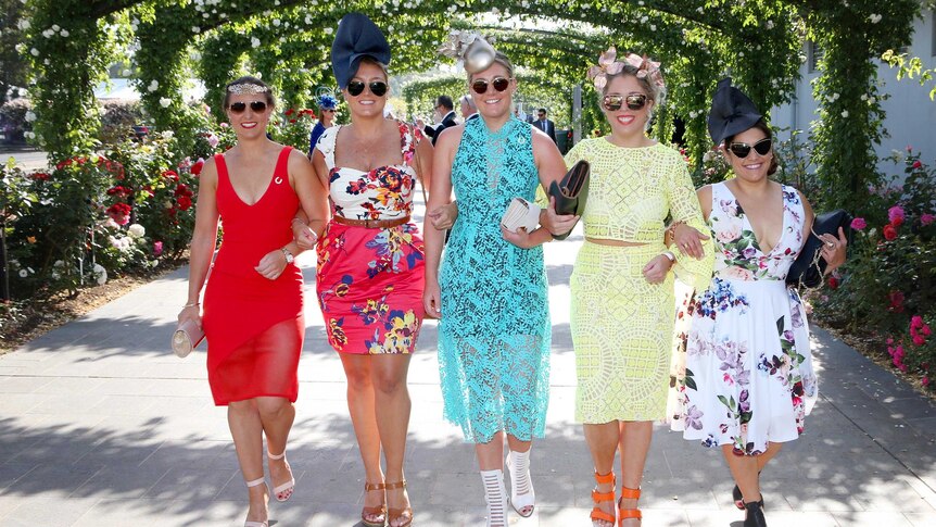 Five women in raceday dresses and hats walk arm-in-arm into Flemington racecourse.