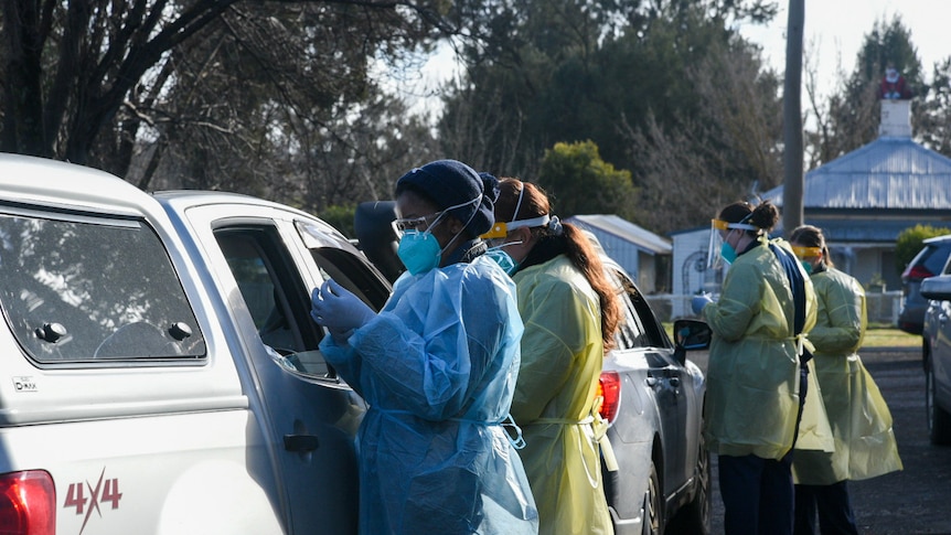 Two nurses in full PPE speak to the occupants of a white ute at a drive-through testing clinic.