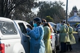 Two nurses in full PPE speak to the occupants of a white ute at a drive-through testing clinic.