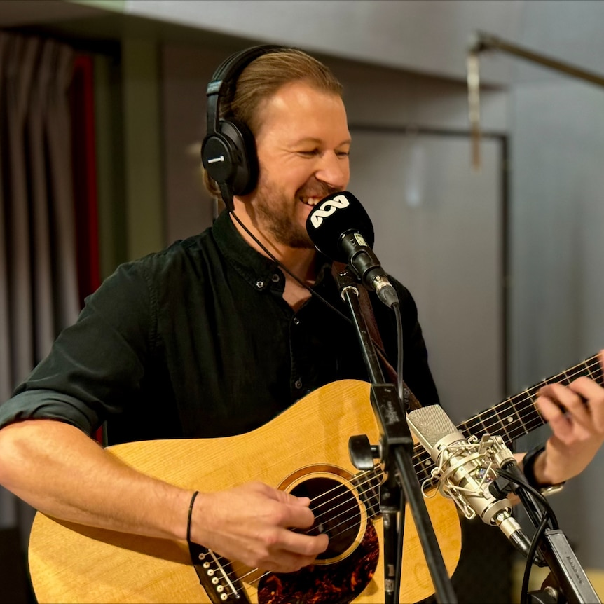 Man with headphones smiles into microphone while playing guitar in a studio.